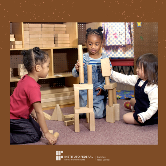 Fotografia com três crianças, sendo uma oriental a direita; uma parda, a esquerda; uma negra, no centro, brincando, numa sala com brinquedos em madeira