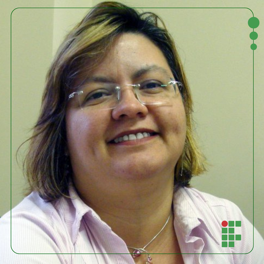 Fotografia de uma mulher sorrindo, de cabelos curtos, usando óculos de lente retangular, sem armação em volta, de brusa branca e usando uma corrente, com pingente, no pescoço